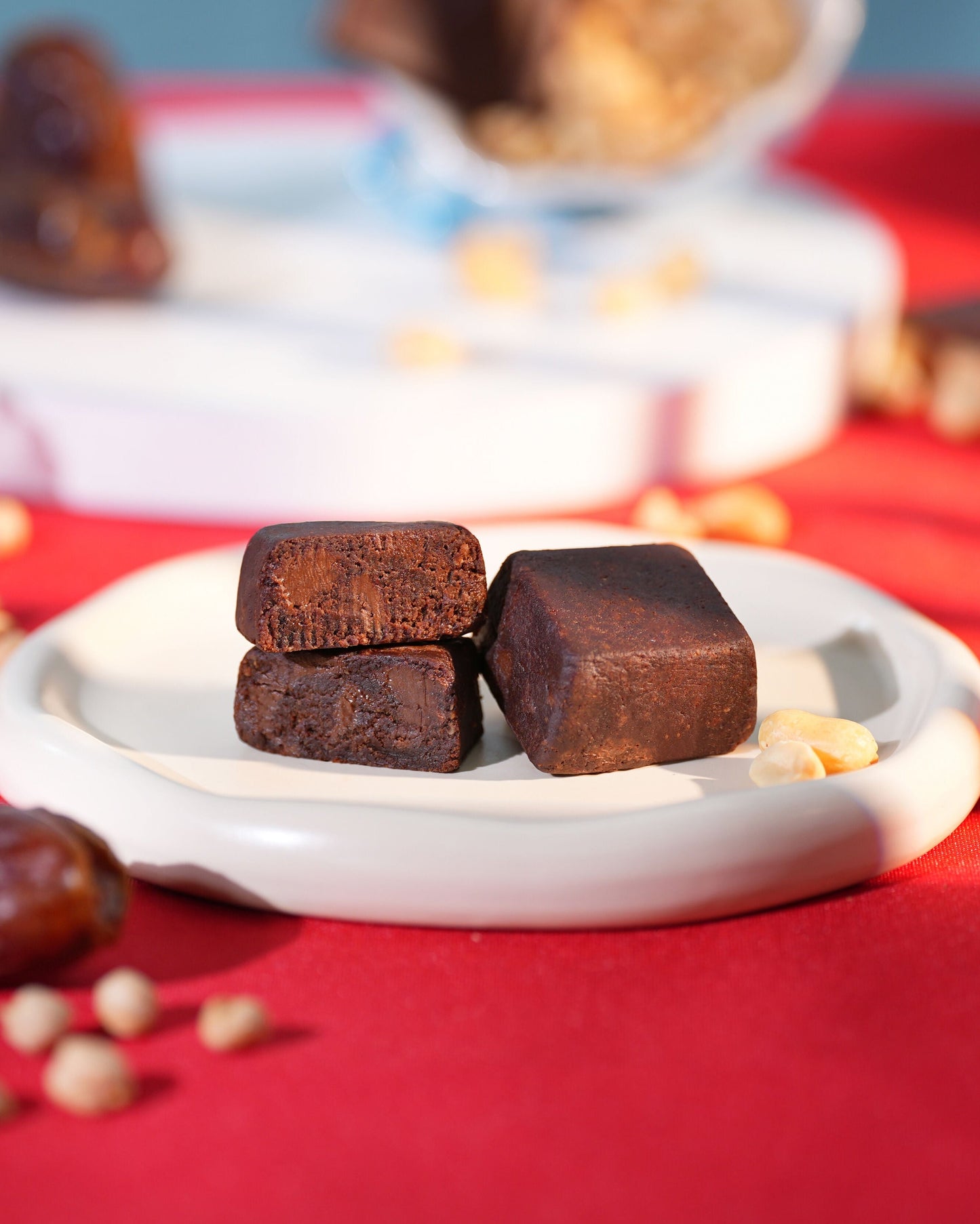 Three dark brown square candies on a white plate with scattered dates and nuts on a red surface.